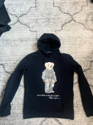 Marinblå  hoodie Polo Bear Ralph Lauren - Snygg svart hoodie från Ralph Lauren med den ikoniska Polo Bear-trycket framtill. Björnen har på sig en cool outfit med skinnjacka och polotröja. Hoodien har dragsko i huvan och ribbade muddar. Perfekt för dig som vill ha en streetstyle-look.