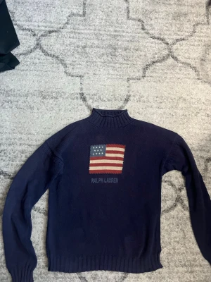 Mörkblå stickad tröja Ralph Lauren - Mörkblå stickad tröja från Ralph Lauren med hög krage och ribbade muddar. På bröstet finns en stor amerikansk flagga i rött, vitt och blått samt broderad text 'RALPH LAUREN' under. Klassisk och stilren design, perfekt till jeans.