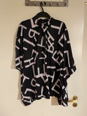 Svartvit blus med grafiskt mönster - Unik svart blus med vitt grafiskt bokstavsmönster och hög krage. Blusen har vid passform och trekvartsärm, vilket ger en avslappnad och trendig look. Perfekt för dig som gillar statement-plagg och vill sticka ut i mängden.