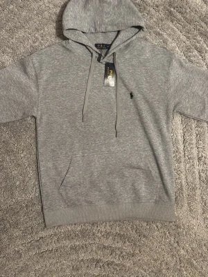 Grå hoodie från Polo Ralph Lauren - Snygg grå hoodie från Polo Ralph Lauren med klassisk huva och dragsnören. Har den ikoniska svarta Polo-loggan broderad på bröstet och en stor magficka framtill. Tillverkad i mjukt material som känns skönt mot huden. Perfekt för en chill och stilren look.
