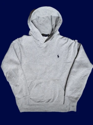 Grå hoodie från Polo Ralph Lauren - Snygg ljusgrå hoodie från Polo Ralph Lauren med klassisk marinblå logga broderad på bröstet. Hoodien har huva, känguruficka och ribbade muddar vid ärmar och nederkant. Tillverkad i mjuk bomullsblandning som är perfekt för chill dagar.