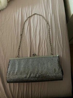 Glittrig silver kuvertväska med kedja - Snygg kuvertväska med glittrig silverframsida och svart baksida. Väskan har en metallkedja som axelrem och klassiskt metallspänne upptill. Perfekt accessoar för att lyfta din outfit med lite extra bling.