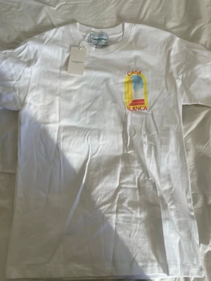 Vit Casa Blanca t-shirt med tryck - Säljer en vit t-shirt från Casablanca med färgglatt tryck av ett fönster och texten 'Casa Blanca' i rött och gult på bröstet och ryggen. Det skrynkliga går att få bort med strykjärn aldrig använd.
