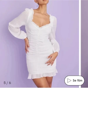 Vit kort volangklänning med v-ringning - Supersöt vit kort klänning med v-ringad hals och volangdetaljer. Klänningen har långa, lätt puffiga ärmar och är gjord i ett tunt, luftigt material. Smickrande rynkningar framtill och volangkant nertill ger en romantisk vibe. Perfekt till studenten. Använd 1 gång, har en fläck på insidan annars fint skick. Liten i storlek