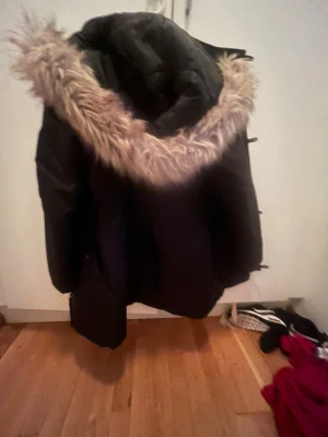 Svart parkasjacka från Only - Snygg svart parkasjacka från Only med stor huva och fejkpäls runt kanten. Jackan har dragkedja och knappar framtill samt flera fickor. Perfekt för kalla dagar och ger en cool streetstil. Materialet är slitstarkt och håller värmen bra.