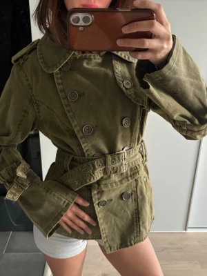 Olivgrön trenchcoat med bälte - Snygg olivgrön trenchcoat med dubbelknäppning, bred krage och bälte i midjan. Jackan har dekorativa axelklaffar och spännen vid ärmsluten för extra stil. Tillverkad i ett robust bomullstyg som ger en cool och avslappnad look. Finns även en drag kedja om man vill ha jackan mer cropped eller längre super najhs. Vintage men super kvalité