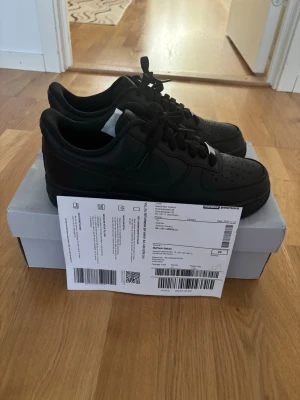 Nike Air Force 1 Low helsvarta sneakers - Säljer ett par klassiska Nike Air Force 1 Low sneakers i helsvart läder. Skorna har en tidlös design med perforerad tå, kraftig sula och ikonisk Swoosh på sidan. Perfekta för dig som vill ha en clean och stilren look.