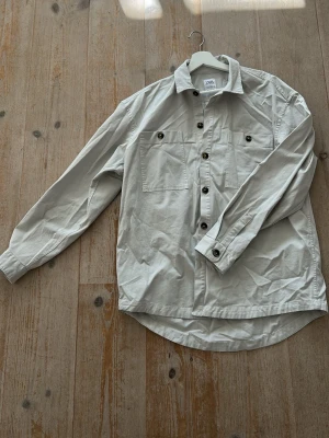 Beige skjorta från Zara med fickor - Stilren beige skjorta från Zara med två bröstfickor och knappar framtill. Skjortan har klassisk krage och långa ärmar med knapp vid manschetten. Perfekt för en avslappnad men snygg look.