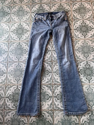 Blå bootcut jeans från True Religion - Säljer ett par ljusblå bootcut jeans från True Religion med klassiska kontrastsömmar och broderade bakfickor. Jeansen har låg midja, fem fickor och detaljerade nitar. Perfekta för dig som gillar en avslappnad men ändå trendig look.