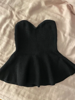 Svart ribbad peplum tubtopp - Säljer en svart ribbad tubtopp med peplumskärning och hjärtformad urringning. Toppen är stickad och sitter snyggt över bysten, perfekt för att skapa en trendig siluett. Supersnygg att styla med jeans eller kjol!