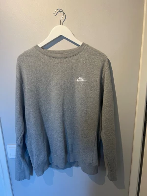 Grå sweatshirt från Nike - Klassisk grå sweatshirt från Nike med rund halsringning och diskret vitt Nike-logotyp broderat på bröstet. Tröjan har långa ärmar och ribbade muddar vid ärmslut och nederkant. 