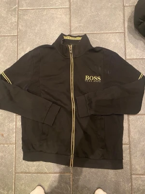 Svart zip hoodie från Hugo Boss - Svart hoodie från Hugo Boss med gulddetaljer och dragkedja framtill. Stort BOSS-tryck på bröstet och ribbade muddar. Snygga gula ränder längs dragkedjan och ärmarna. Tillverkad i mjukt bomullsmaterial för en bekväm känsla.