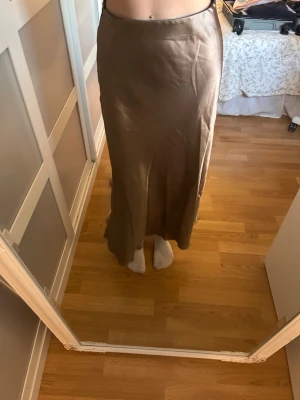 Beige långkjol i satin - Säljer en beige långkjol i mjukt satinliknande material. Kjolen har en enkel och stilren design med lätt utsvängd form som ger en snygg siluett. Perfekt att matcha med crop tops eller stickade tröjor för en trendig look.