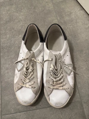 Vita Philippe Model sneakers med grå detaljer - Snygga vita sneakers från Philippe Model med grå och silverfärgade detaljer samt svart hälparti. Skorna har text på skosnörena och en unik paneldesign i mocka och läder. Perfekta för dig som gillar streetstyle och vill ha något som sticker ut. Defekter på snörena så som knutar osv