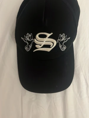 Svart keps med broderad S och duvor - Snygg svart keps med stor vit broderad S på framsidan och två broderade duvor på sidorna. Justerbar rem baktill för perfekt passform. Kepsen har klassisk böjd skärm och är tillverkad i ett mjukt tygmaterial.