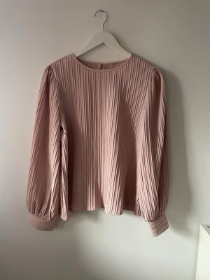 Plisserad rosa blus med långa ärmar - Plisserad blus i ljusrosa med rund halsringning och långa puffärmar med breda muddar. Blusen har en knappdetalj i nacken och är tillverkad i ett lätt, flowy tyg.