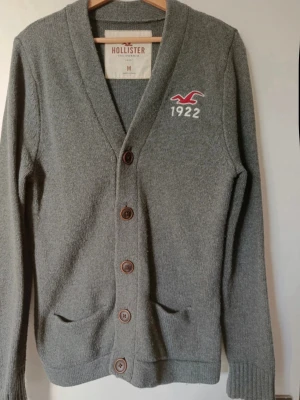 Grå cardigan från Hollister - Snygg grå kofta från Hollister med knappar framtill och två fickor. Broderad logga och siffran 1922 på bröstet. Mjuk och skön stickad känsla, perfekt för lager-på-lager. Klassisk design med ribbade muddar och v-ringad hals.