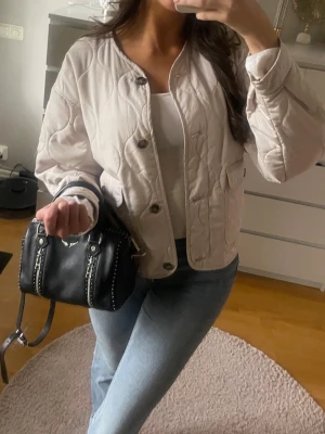 Beige quiltad jacka - Snygg beige quiltad cardiganjacka med rund halsringning och stora knappar framtill. Jackan har två framfickor och en avslappnad passform. Perfekt att slänga över en topp för en trendig look.