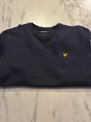 Mörkblå sweatshirt från Lyle & Scott - Klassisk mörkblå sweatshirt från Lyle & Scott med rund halsringning och gul broderad logotyp på bröstet. Tröjan har ribbade muddar vid ärmslut och nederkant. Perfekt för en avslappnad och stilren look. Pris kan diskuteras!