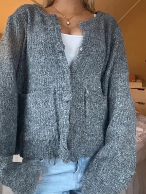 Grå stickad kofta med fickor - Mysig grå stickad kofta med två framfickor och knappar framtill. Koftan har en avslappnad passform, långa ärmar och är tillverkad i ett mjukt, fluffigt material. Perfekt att slänga på sig över en t-shirt eller linne.