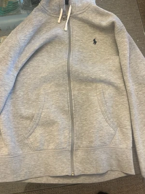 Grå hoodie från Polo Ralph Lauren - Snygg grå hoodie från Polo Ralph Lauren med dragkedja framtill, två fickor och klassisk huva med vita snören. Liten mörkblå logga broderad på bröstet. Tillverkad i mjuk bomullsblandning som känns skön mot huden. Perfekt för en chill och stilren look.