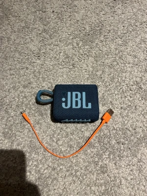 Blå JBL Go 3 bärbar högtalare - Säljer en blå JBL Go 3 bärbar högtalare med robust tygmaterial och integrerad ögla för enkel upphängning. Kommer med en orange USB-laddningskabel. Smidig och kompakt design, perfekt för att ta med överallt.