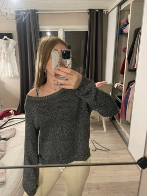 Mörkgrå stickad tröja med bred hals - En mörkgrå stickad tröja med bred, lite off-shoulder halsringning och långa ärmar. Tröjan har ribbstickade muddar vid ärmslut och nederkant. Perfekt för att skapa en avslappnad och trendig look.