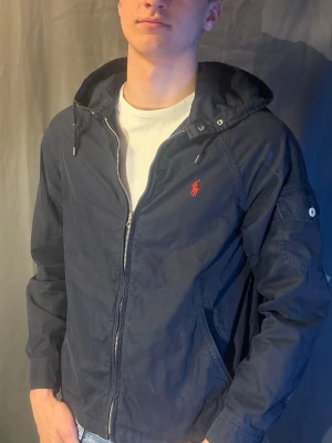 Marinblå vindjacka från Polo Ralph Lauren - Snygg marinblå vindjacka från Polo Ralph Lauren med dragkedja framtill, huva med dragsko och röd logga på bröstet. Jackan har fickor med dragkedja och knappdetalj på ärmen. Modellen är 190cm och väger 80kg. Skriv om du har frågor 🥰
