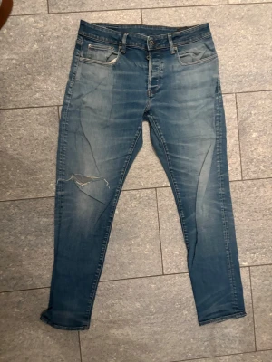 Blå slitna slim 3301  jeans med hål från Raw - Säljer ett par blå skinny jeans med slitningar och ett stort hål på vänster ben. Jeansen har klassisk femficksdesign, dragkedjegylf och är i stretchigt denimtyg. Perfekta för en avslappnad och trendig look.