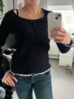 Svart ribbad topp med knappar - Svart långärmad ribbad topp med tre knappar framtill och vit volangkant längst ner. Är i bra skick i storlek S