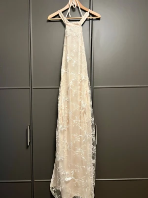 Lång ljus klänning med spetsdetaljer - Elegant långklänning i ljus beige med tunna axelband och vacker spetsbrodyr i vitt. Klänningen har ett transparent lager med blommigt mönster och en mjuk, flytande siluett. Perfekt för speciella tillfällen när du vill känna dig extra fin.