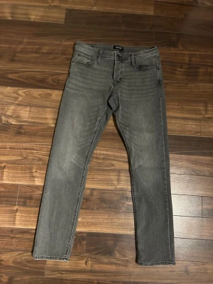 Grå Jack and Jones slim fit jeans - Säljer ett par grå slim fit jeans med klassisk femficksdesign och diskreta sömmar. Jeansen har en något tvättad look och raka ben. Passar perfekt till en avslappnad stil och är tillverkade i mjukt denimtyg för skön komfort.