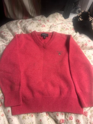 Rosa V-ringad tröja från GANT - Rosa stickad tröja från GANT med V-ringning och diskret logga broderad på bröstet. Tröjan har ribbade muddar vid ärmslut och nederkant, vilket ger en klassisk och stilren look. Perfekt för dig som gillar tidlös design och mjuka material.