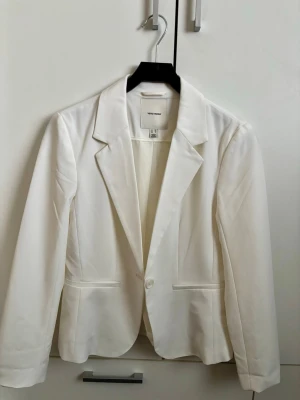 Vit kavaj från Vero Moda - Stilren vit kavaj från Vero Moda med klassisk krage och en knapp framtill. Kavajen har två fickor och är helfodrad för en snygg passform. Perfekt för att lyfta vilken outfit som helst med en fräsch och modern look.