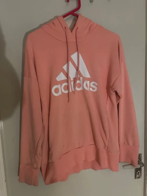 Adidas rosa hoodie med logga - Säljer en snygg rosa hoodie från Adidas med stor vit logga på bröstet. Hoodien har huva med dragsko och breda muddar vid ärmslut och nederkant. Perfekt för en avslappnad och sportig stil. Materialet är mjukt och skönt, troligtvis bomullsblandning.