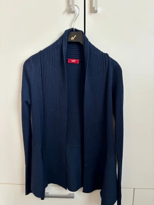 Mörkblå ribbad kofta från Esprit - Snygg mörkblå kofta från Esprit med öppen front och ribbad struktur. Långärmad modell med asymmetrisk nederkant och mjukt, stretchigt material. Perfekt för lager på lager och enkel att matcha med olika outfits.