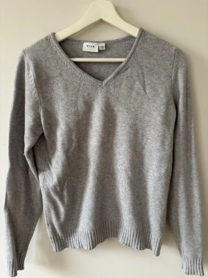 Grå stickad tröja från Vila - Mysig grå stickad tröja från Vila med v-ringning och långa ärmar. Tröjan har ribbstickade muddar vid ärmslut och nederkant, vilket ger en snygg och enkel look. Perfekt för lager på lager och passar till det mesta. Skön material. 