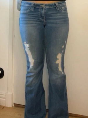 Blå bootcut jeans med slitningar - Säljer ett par ljusblå bootcut jeans med slitna detaljer framtill och dekorativa bakfickor med knappar. Jeansen har klassisk femficksmodell och normal midja. Perfekta för en avslappnad och trendig look.
