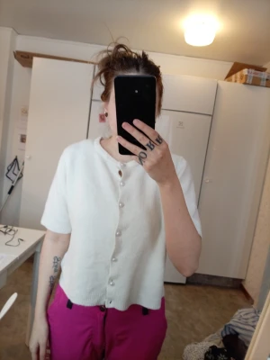 Vit kortärmad kofta med pärlknappar - Säljer en vit kofta med korta ärmar och pärlknappar framtill. Koftan har en rund halsringning och är tillverkad i ett mjukt stickat material. Perfekt att slänga över en t-shirt eller bära som den är. (ena pärlan längst ner ä av) 