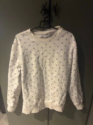 Vit sweatshirt från Calvin Klein Jeans 13-15år - Vit sweatshirt från Calvin Klein Jeans med blått CK-mönster över hela tröjan. Klassisk rund halsringning och långa ärmar. Tillverkad i mjuk bomull för en bekväm känsla och avslappnad passform.