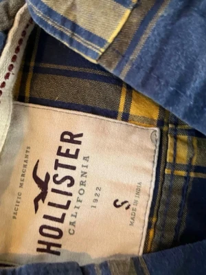 Rutig skjorta från Hollister blå/gul - Säljer en snygg rutig skjorta från Hollister i marinblått och gult. Skjortan har klassisk krage, knappstängning framtill och en bröstficka med Hollisters logga broderad. Tillverkad i mjuk bomull, perfekt för en avslappnad stil.