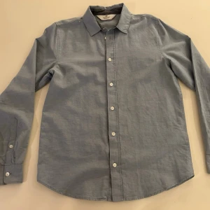 Ljusblå skjorta med knappar - Klassisk ljusblå skjorta från H&M med lång ärm och vita knappar framtill. Skjortan har en rak passform och är tillverkad i mjuk bomull, perfekt för en clean och enkel stil.