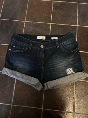Mörkblå denimshorts med uppvik - Snygga mörkblå denimshorts från George med uppvikta ben och klassiska femficksdetaljer. Shortsen har en liten slitning på ena benet för en cool look och är tillverkade i bomull. Perfekta för sommarens alla äventyr.