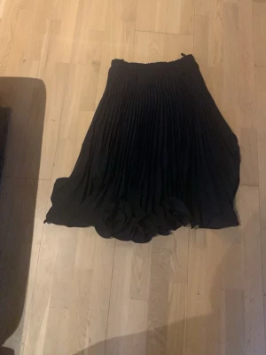 Svart plisserad midikjol - Säljer en svart plisserad midikjol med elastisk midja. Kjolen har ett snyggt fall och är gjord i ett lätt, flytande tyg som ger en elegant look. Perfekt att matcha med både sneakers och boots för en trendig stil.