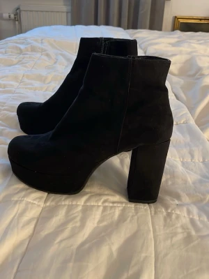 Svarta stövletter med klack - Säljer ett par svarta stövletter i mockaimitation med hög, bred klack och platåsula. Skorna har en stilren siluett och dragkedja på insidan för enkel påtagning. Perfekta för dig som vill ha ett par trendiga boots med lite extra höjd.