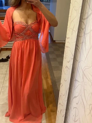 korallfärgad lång balklänning - Säljer en korallfärgad lång balklänning från Cotton Club med axelbandslös design och glittriga stenar över bysten. Klänningen har markerad midja och ett flytande, elegant fall. Perfekt för bal eller festliga tillfällen. Jag som bär den är vanligtvis en storlek s så skulle gissa på att den är kring den storleken! Går även att lägga till band om man känner för det för det finns möjlighet på det i insidan av klänningen men har inga band! Man får även med den matchande sjalen på första bilden!!