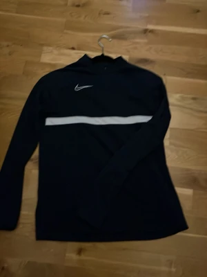 Svart långärmad träningströja Nike - Svart långärmad träningströja från Nike med vit logga på bröstet och en vit horisontell rand över bröstet. Perfekt för träning eller sportaktiviteter. Tröjan har en sportig design och är tillverkad i ett lätt och bekvämt material.
