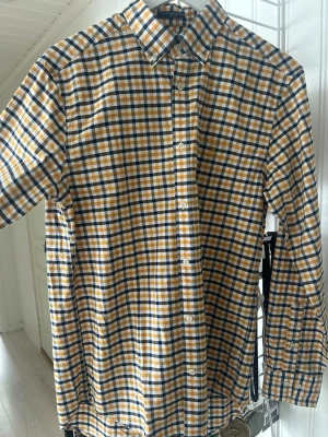Rutig skjorta från Gant i bomull - Oanvänd Klassisk långärmad skjorta från Gant med rutigt mönster i vitt, blått och gult. Skjortan har knappar framtill, bröstficka med broderad logga och en bekväm passform. Perfekt för dig som gillar stilrena och tidlösa plagg. Säljer då jag inte fått användning av plagget.. 