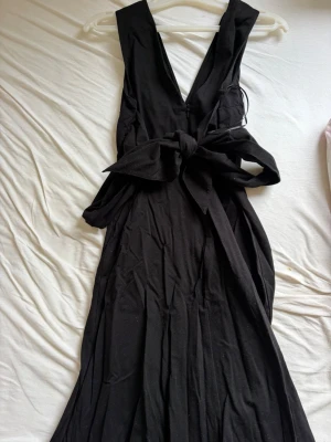 Svart midi klänning ifrån Massimo dutti passar xs-s - Elegant svart långklänning med djup v-ringning och breda axelband. Klänningen har en stor dekorativ rosett i midjan och är tillverkad i ett mjukt tyg som faller snyggt. Perfekt för dig som vill ha en stilren och klassisk look.