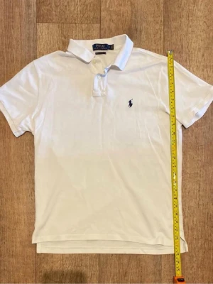 Vit pikétröja från Polo Ralph Lauren - Klassisk vit pikétröja från Polo Ralph Lauren med korta ärmar och blå broderad logga på bröstet. Tröjan har krage och knappar framtill. Perfekt till en avslappnad och stilren look.